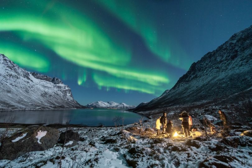 People standing around Camp fire with Northern lights over them