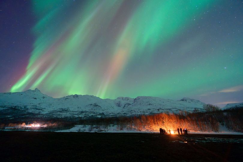 People standing around bonfire with Northern lights over them