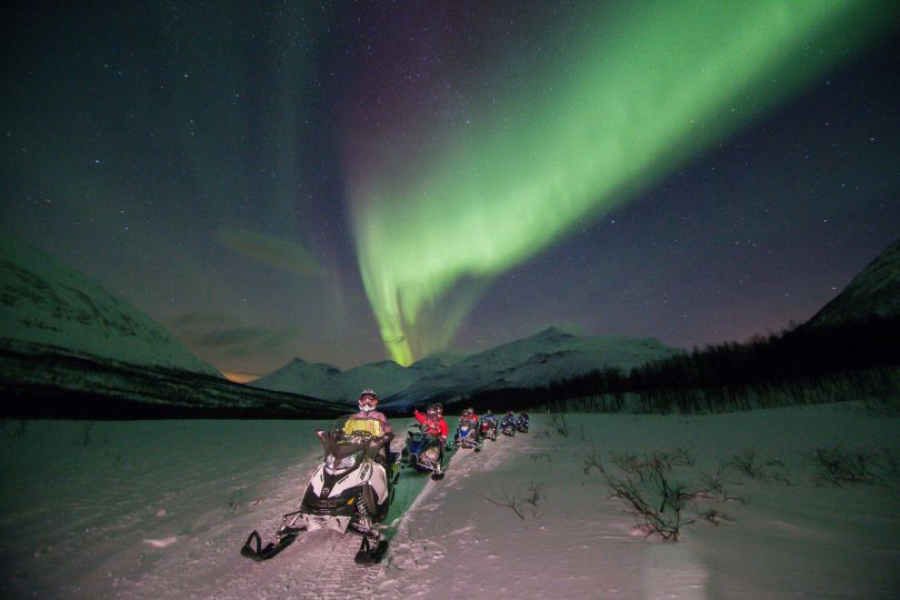 People driving snowmobiles through snowy nature under Northern lights